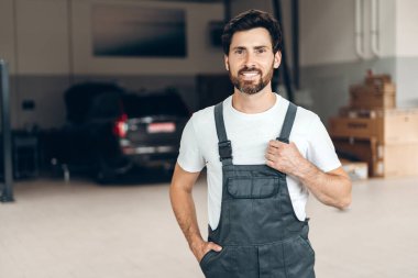 Profesyonel tamirci oto tamirhanesinde gururla duruyor, tulum giyiyor ve gülümsüyor, arka planda bir araba var, uzmanlık ve otomotiv hizmetlerini temsil ediyor.