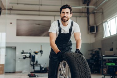 Siyah eldiven ve tulum giyen gülümseyen tamirci bir oto tamirhanesinde araba lastiği tutuyor.