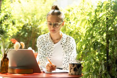 Gözlük takan genç bir kadın dizüstü bilgisayarıyla uzaktan çalışırken notlar alıyor. Huzurlu bir bahçe ortamının temiz havasının ve doğal ışığının tadını çıkarıyor. Eğitim kavramı