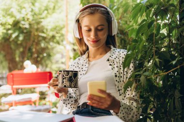 Serbest çalışan genç kadın kahve içiyor, kulaklıkla müzik dinliyor ve teneffüste akıllı telefon kullanıyor, bahçede defter ve kalemle masada oturuyor.