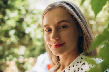 Uzun açık kahverengi saçlı ve çarpıcı kırmızı rujlu genç bir kadın canlı bir bahçede gülümsüyor, siyah benekli şık bir beyaz gömlek giyiyor, mutluluk ve cazibe saçıyor.