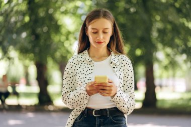 Gündüz giyimli genç bir kadın güneşli bir yaz gününde parkta duruyor ve akıllı telefonunu kullanıyor, sosyal medyada geziniyor ve çevrimiçi iletişim, sosyal medya kavramının tadını çıkarıyor.