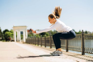 Nehir kenarındaki bir parkta zıplayan konsantre sporcu kadın, güneşli yaz gününde uzun kollu beyaz gömlek ve siyah tayt giyerek egzersiz yapıyor. Egzersiz konsepti