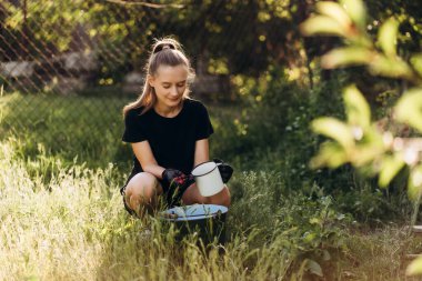 Eldiven takan genç bir kadın, neşeli bir yaz günü güneşli bahçesinde yemyeşil yeşilliklerle çevriliyken metal bir kupada taze organik böğürtlen topluyor.
