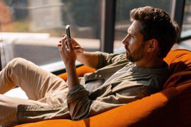 Genç işadamı, modern ofisin penceresinin yanındaki rahat turuncu puf minder üzerinde dinlenirken akıllı telefon kullanıyor.