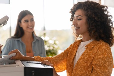 Modern bir ofiste çalışan iş kadınları, doğal ışık ile çevrili çok işlevli bir yazıcı kullanarak verimli bir şekilde kopyalarını çıkarıyorlar.