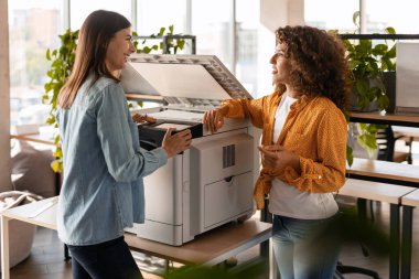 İki kadın ofis çalışanı, çok işlevli bir yazıcıya bağlanıyor. Belgeleri tartışıyor ve modern bir iş yerinde gülücüklerini paylaşıyorlar.