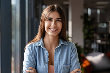 Modern bir ofiste kollarını kavuşturarak gülümseyen neşeli bir iş kadınının portresi güven ve profesyonellik sergiliyor.