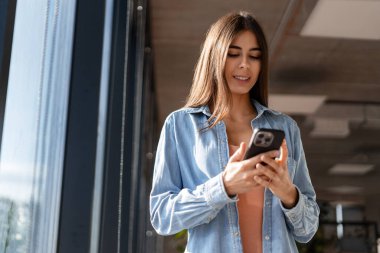 Gülümseyen iş kadını cep telefonu kullanıyor, ofiste tatilin tadını çıkarıyor, pencerenin yanında duruyor.