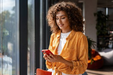 Güneşli bir günde modern ofiste pencerenin yanında cep telefonu kullanan gülümseyen bir iş kadını.