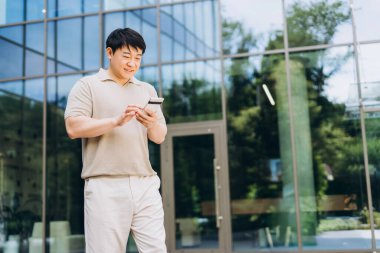 Asyalı işadamı, akıllı telefonu ve kredi kartıyla online ödeme yaparken gülümsüyor. Modern ofis binasının önünde cam cepheli duruyor. Çevrimiçi teknoloji kavramı