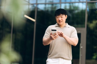 Asyalı adam, modern bir cam binanın önünde modern teknolojinin rahatlığını sergilerken online alışveriş ve mobil bankacılık için akıllı telefonunu ve kredi kartını kullanıyor.