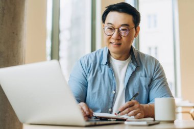 Asyalı serbest çalışan gülümseyen ve dizüstü bilgisayar üzerinde çalışırken not alan, uzaktan çalışma ve esnek programdan hoşlanan biri. Çevrimiçi eğitim kavramı