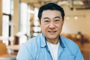 Kendinden emin bir şekilde gülümseyen, orta yaşlı Asyalı bir adam, çağdaş ofis ortamında profesyonelliği ve başarıyı somutlaştırıyor, sıcaklığını ve yaklaşımını yansıtıyor.