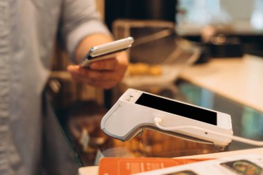 Müşteri akıllı telefonu pos terminalinin üzerinde tutuyor, modern mağazada bağlantısız ödeme yapıyor, hızlı ve güvenli işlemler için NFC teknolojisinden faydalanıyor.