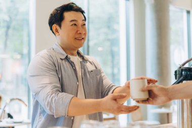 Neşeli Asyalı bir adam bir meslektaşından bir fincan kahve alır, modern ofis ortamlarında bir ara verdiklerinde dostluk ve iletişim duygusunu teşvik eder.