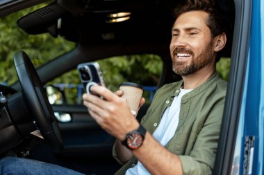Gülümseyen işadamı arabasında oturuyor, cep telefonunu kullanırken kahvenin tadını çıkarıyor, sabah işe gidip gelirken modern teknolojiyi kucaklıyor.