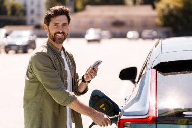 Gülümseyen adam, şehrin hareketli otoparkında akıllı telefon uygulamasını kullanırken elektrikli arabasını şarj ediyor, modern, çevre dostu teknolojiyi kucaklıyor.