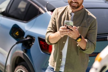 Bir şarj istasyonunda elektrikli arabayı şarj ederken cep telefonuyla meşgul olan gülümseyen adam, sürdürülebilir ulaşım çözümlerini kucaklıyor