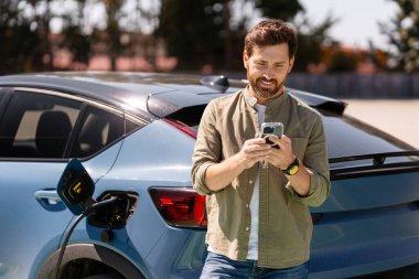 İş adamı akıllı telefonuyla meşgul olurken şarj istasyonunda elektrikli bir arabayı şarj ediyor, sürdürülebilir ulaşım ve temiz enerjiyi vurguluyor