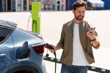 Gülümseyen adam şarj kablosu tutuyor ve şarj istasyonunda elektrikli araba şarj ederken cep telefonu kullanıyor.