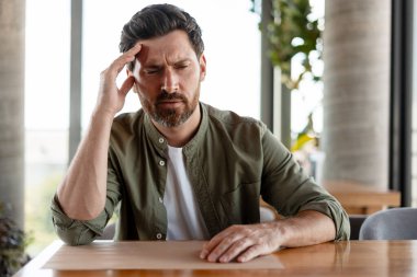 Stresli iş adamı, yoğun bir kafede uzaktan çalışırken, baş ağrısı ve yorgunlukla boğuşuyor.
