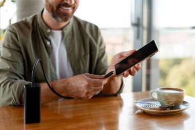 Gülümseyen işadamı kahvesini içerken bir kafedeki akıllı telefonu güç bankasına bağlıyor, işi ve rahatlığı modern ortamlarda harmanlıyor.