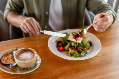 Çatal ve bıçak kullanan, feta peynirli, domatesli, zeytinli ve marullu sağlıklı Yunan salatası yiyen adama yaklaş.