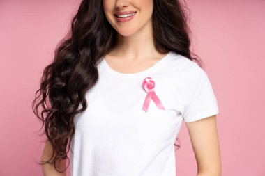 Beyaz tişörtlü gülümseyen kadın pembe kurdeleyle süslenmiş, göğüs kanseri farkındalığını gururla desteklerken canlı pembe bir arka plan, pozitif enerji ve umut yayıyor.