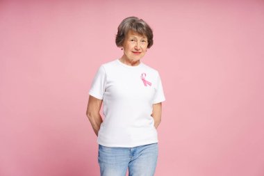 Beyaz tişörtlü, pembe kurdeleli mutlu olgun kadın pembe arka planda poz veriyor, göğüs kanseri farkındalığı kampanyasını destekliyor, umut ve dayanışmayı ifade ediyor. Destek konsepti