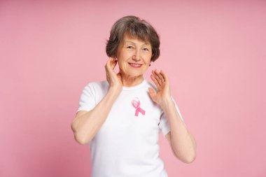 Pembe kurdeleyle süslenmiş beyaz tişört giyen mutlu bir son sınıf öğrencisi, canlı pembe arka planda poz verirken göğüs kanseri farkındalığını gururla destekliyor. Sağlık hizmeti kavramı