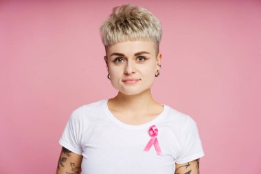 Beyaz tişörtüne pembe kurdele takmış kısa sarı saçlı ve dövmeli genç bir kadının stüdyo portresi meme kanseri farkındalığını sembolize ediyor. Destek konsepti