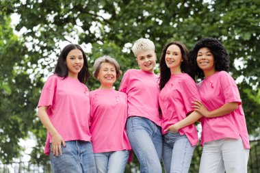 Bir grup çok uluslu gülümseyen kadın pembe tişörtler giyip göğüs kanseri farkındalığını destekliyor kameraya bakıyor, sokakta poz veriyorlar. Tedavi kavramı