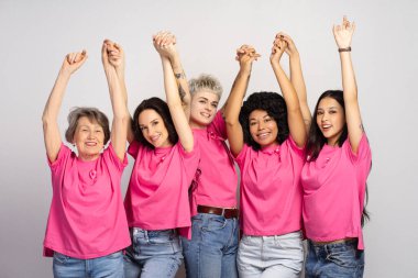 Çeşitli gülümseyen kadınlar el ele tutuşup göğüs kanseri farkındalığı için kollarını kaldırarak kameraya bakıyorlar. Sağlık hizmeti kavramı