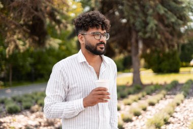 Yeşil bir parkta kahve molası veren genç bir işadamı, elinde tek kullanımlık bir bardakla başka yere bakıyor.