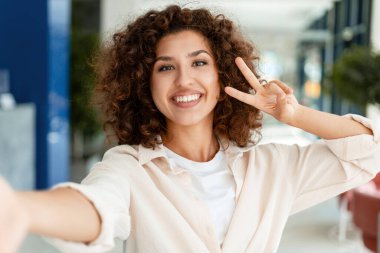 Kıvırcık saçlı, gülümseyen genç bir kadının portresi. Selfie çekiyor ve zafer işaretini parmaklarıyla yapıyor. Neşe ve pozitifliği ifade ediyor. Eğlenceli konsept