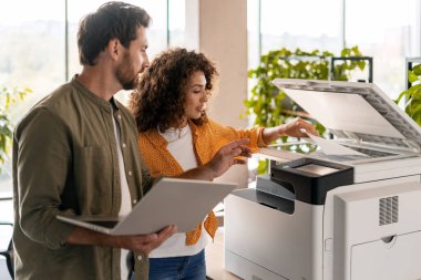 Bir ofiste iş birliği yapan iki iş arkadaşı, biri dizüstü bilgisayarı yönetirken, diğeri de yazıcı ayarlarını ince ayar yaparak, takım çalışmasını ve modern teknolojiyi iş başında gösteriyor.