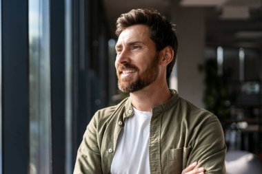 Mutlu girişimci gündelik giysiler giyer kolları çapraz duran, gülümseyen ve gelecekteki başarılarını düşünen modern ofis ortamında doğal ışığın tadını çıkaran. İş konsepti