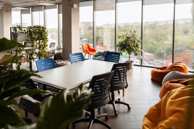 Siyah ve mavi sandalyeli büyük beyaz bir masa, puf minderleri ve çeşitli bitkilerin oluşturduğu ortak ofis alanı rahatlatıcı ve üretken bir ortam yaratır.