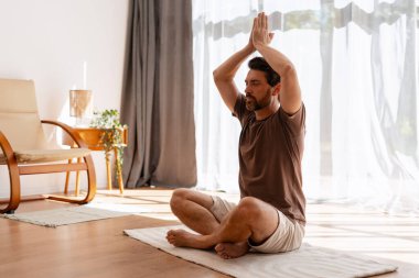Orta yaşlı bir adam parlak bir odada bacak bacak üstüne atarak oturur, yoga meditasyonunu ellerini başının üstüne kaldırarak yapar, farkındalığı, dengeyi ve iç huzuru kucaklar.
