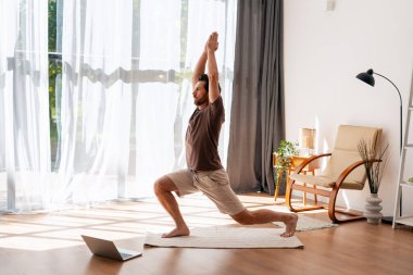 Sakallı, orta yaşlı, parlak modern oturma odasında yoga yapan bir adam, online bir dersi takip ediyor. Çevrimiçi teknoloji kavramı