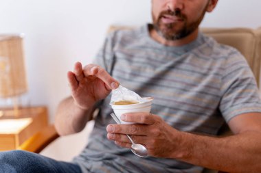 Sakallı, orta yaşlı bir adam evde seçici odak noktasında yoğurt kapağını açarken rahatlıyor. Sağlıklı beslenme kavramı
