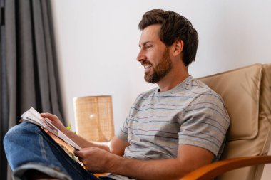 Rahatlamış sakallı Latin adam evde oturmuş modern oturma odasında kitap okurken keyfine bakıyor. Hobi, rahatlatıcı konsept