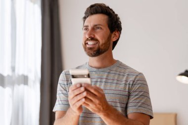 Modern oturma odasında gülümserken akıllı telefon tutan mutlu, sakallı bir Latin adam. Çevrimiçi teknoloji kavramı