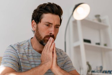 Düşünceli bir adam, elleri birbirine bastırılmış, düşünceli bir şekilde aşağıya bakıyor, yumuşak doğal ışıkla aydınlık, modern bir iç mekanda oturuyor ve bir yansıtıcı yaratıyor. Kötü haber konsepti