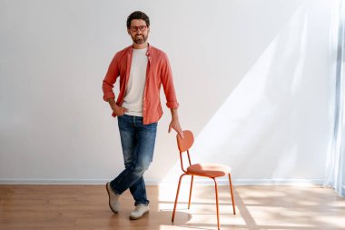 Gündelik kıyafetli kendine güvenen Latin bir adam modern sandalyeyle içeride dikiliyor. Yakışıklı manken pozu, modern stil oturma odasında duruyor. Reklam kavramı