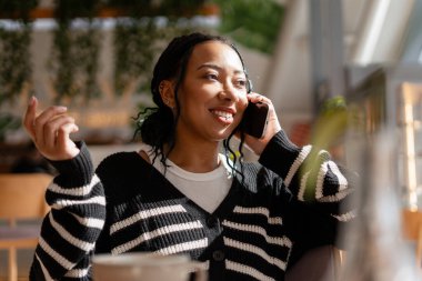Gülümseyen genç siyahi kadın telefon görüşmesi yapıyor, modern bir kafe ortamında iletişim kuruyor ve iletişim kuruyor.