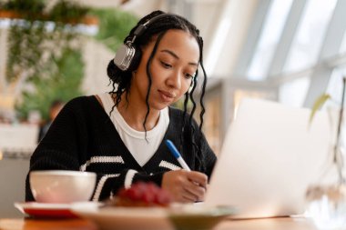 Örgü ören, dizüstü bilgisayar kullanan ve kafede kulaklık takan gülümseyen genç bir kadın.