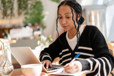 Kahve dükkanında örgü ören, dizüstü bilgisayara yazı yazan, odaklanmış ve profesyonel olan genç melez kadın.