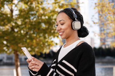 Rasta saçlı, kulaklıkla müzik dinleyen ve parkta akıllı telefonuyla sosyal medyayı kontrol eden mutlu genç bir kadın.
